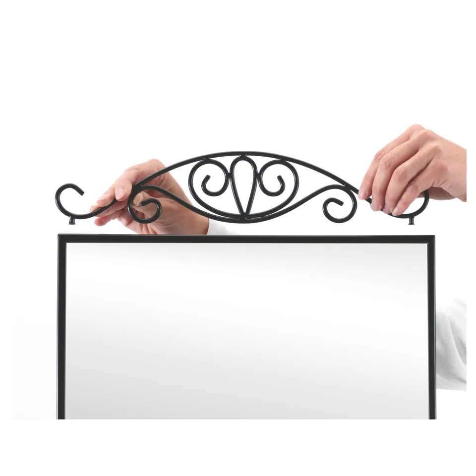 Two hands adjust a decorative metal crown atop a rectangular mirror. The crowns scroll design adds elegance.