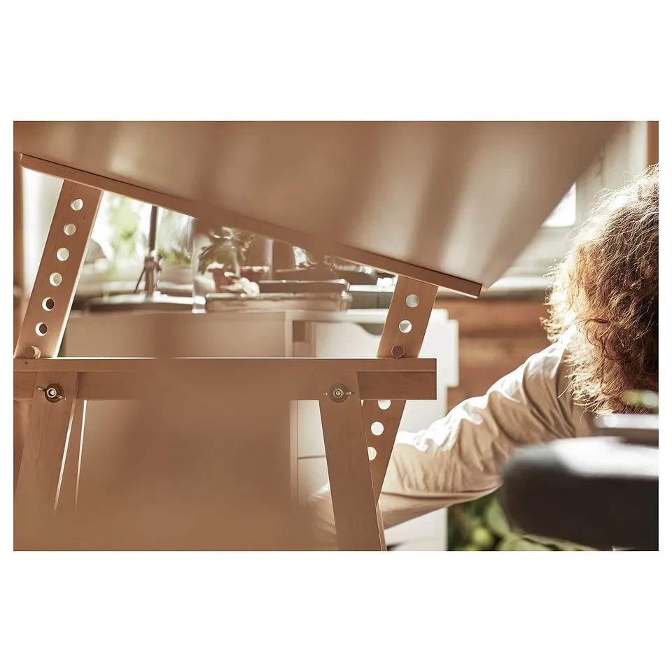 A person adjusting a wooden LAGKAPTENs desk. The desk has a light wooden top and slanted trestles. Its set up indoors with natural lighting.
