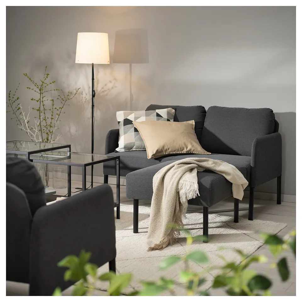 Cosy living room with GLOSTAD grey sofa, black coffee table, and floor lamp. Neutral pillows and throw blanket on sofa. Plant and rug on wooden floor.