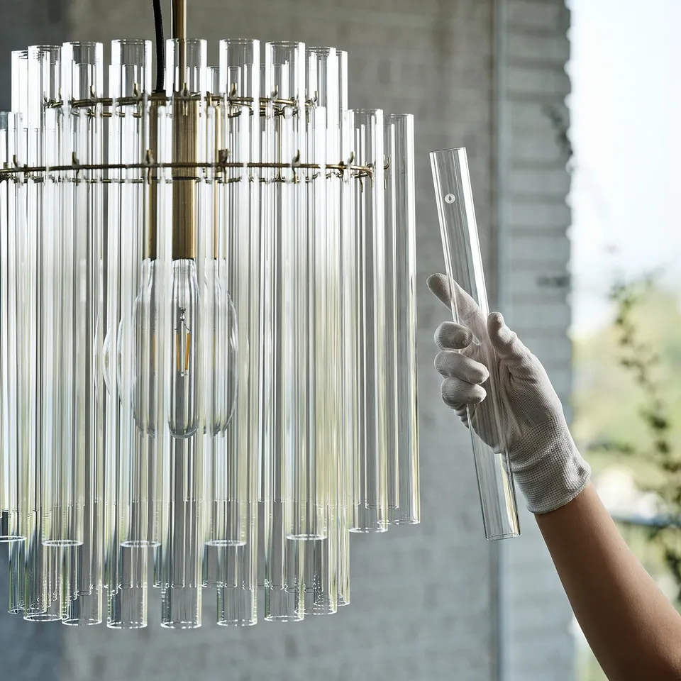 Gloved hand adjusts brass chandelier with glass tubes by large window.