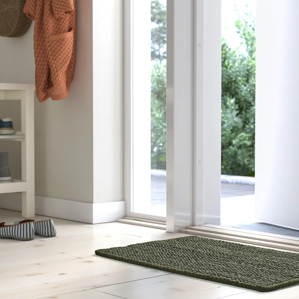 Cosy entryway with LANDBANA dark green jute mat, striped slippers, hanging sweater, and white floors. Mat size similar to two slippers.