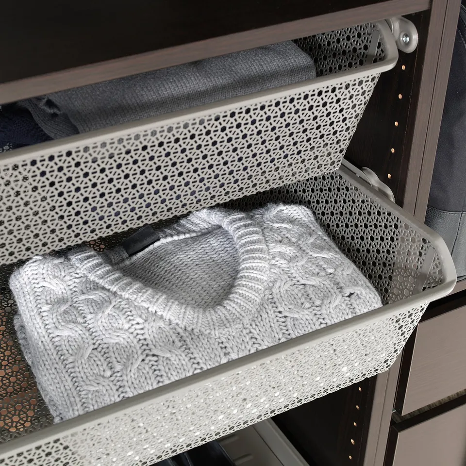 A metal mesh basket in a wardrobe, holding folded clothes, part of IKEAs KOMPLEMENT organisers.