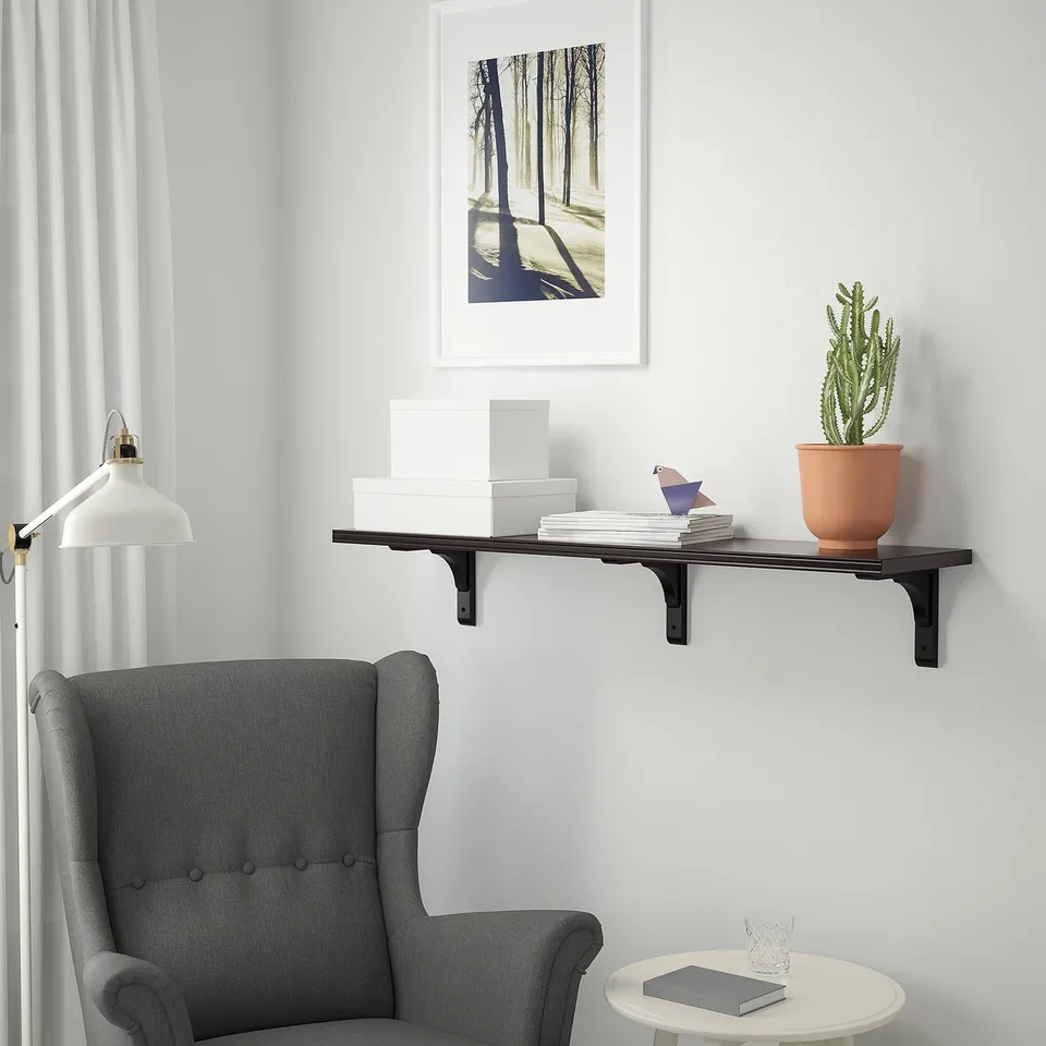 Minimalist wall shelf BERGSHULT holds books, decor near grey chair. Framed forest art above, white lamp left, cactus right.