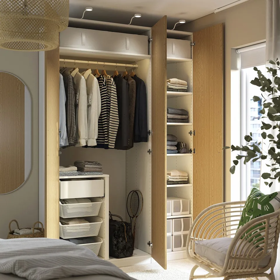 Modern open wardrobe with clothes, lighting, and drawers near bed and window.