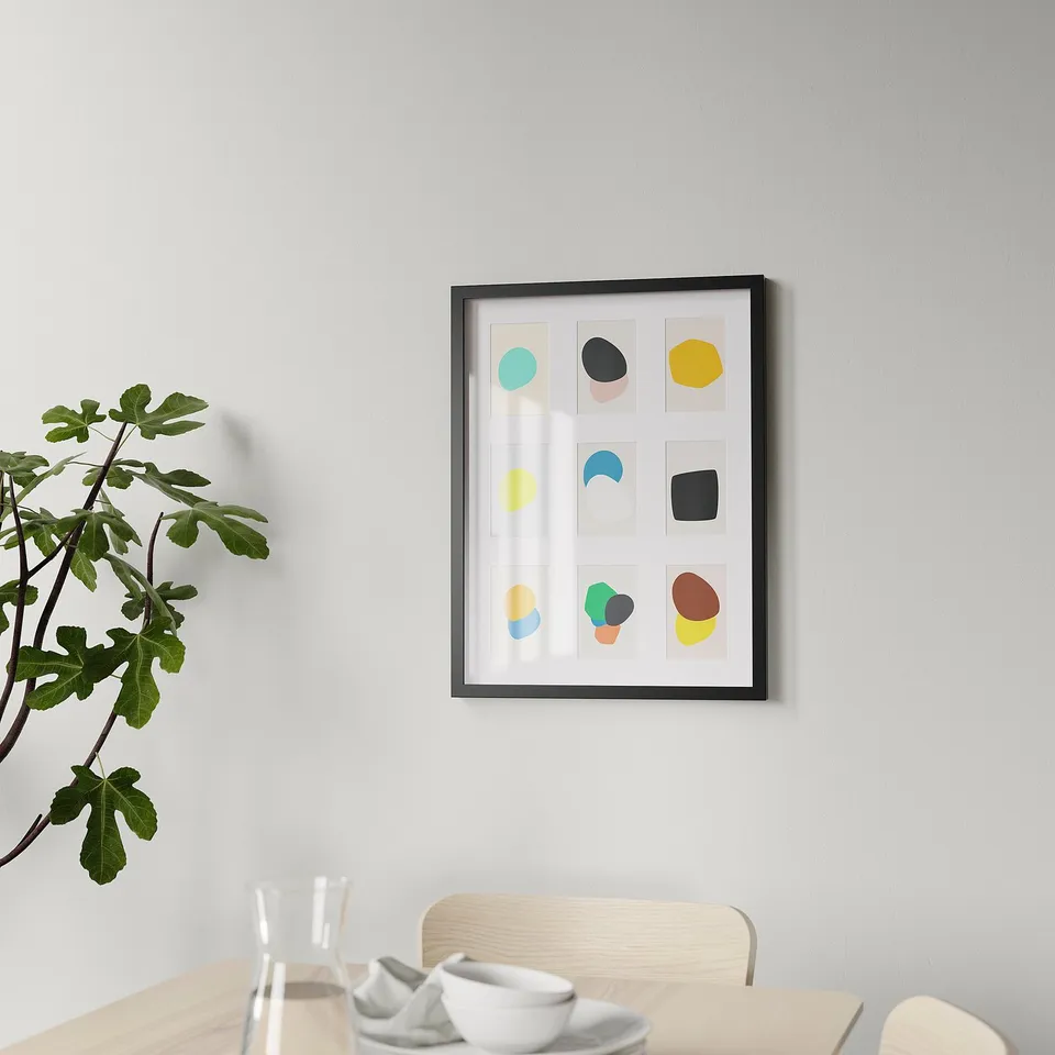 Modern photo frame with abstract art hangs on wall over dining table with dishes and water. Nearby, a potted plant adds greenery.