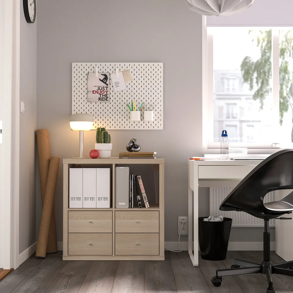 Home office with KALLAX storage unit, black chair, white desk by window, lamp, and pegboard.