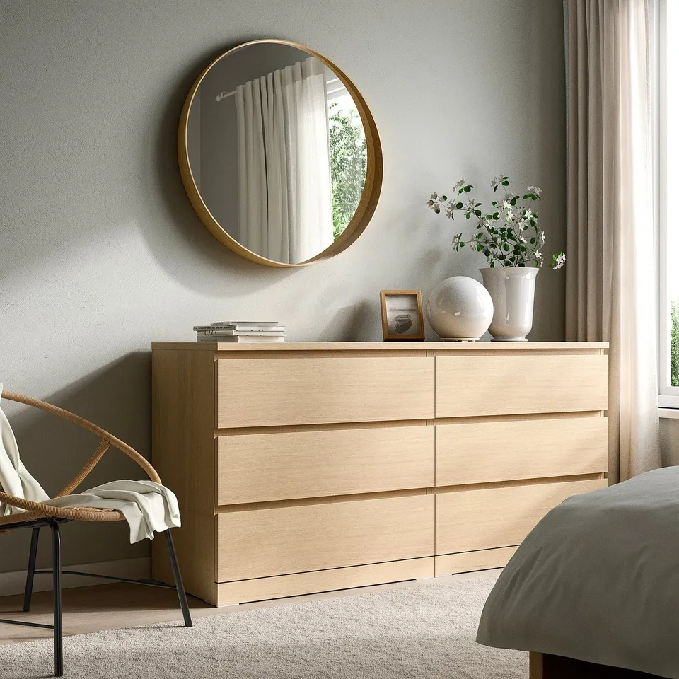 Wooden dresser with 6 drawers, large gold mirror, vases, books, and frame. Wicker chair with blanket, bed with grey comforter.