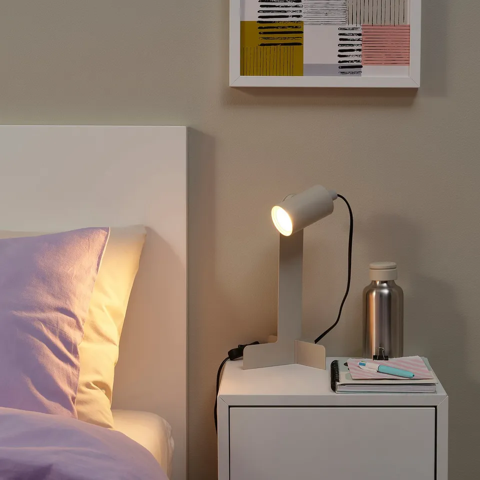 Small white FLOTTILJ lamp on bedside table next to bed, illuminating a cosy space.