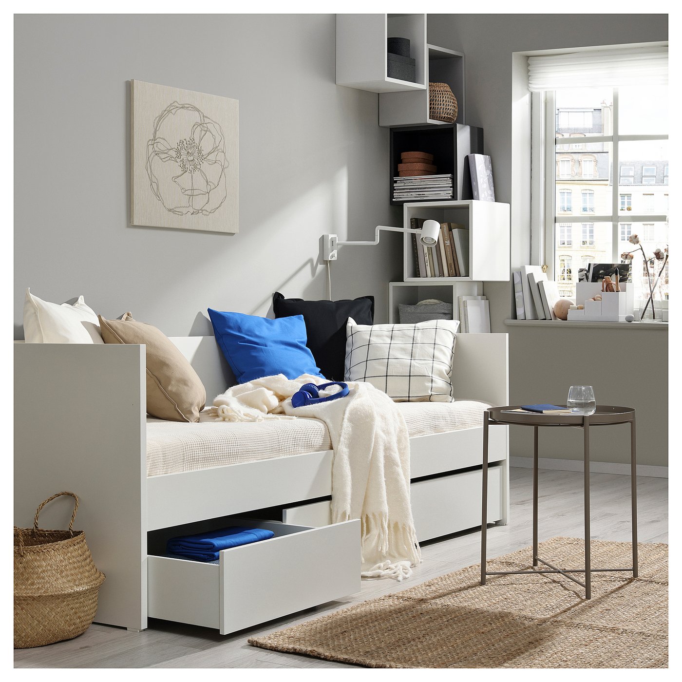Modern bedroom with white VIHALS daybed, colourful pillows, shelf unit, woven rug, and small table by a window.