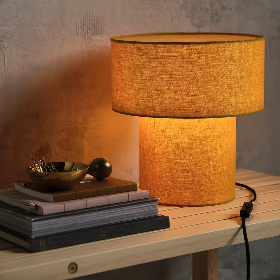 A lit table lamp on a wooden shelf emits warm light, beside books and decor, enhancing the cosy stockholm collection atmosphere.