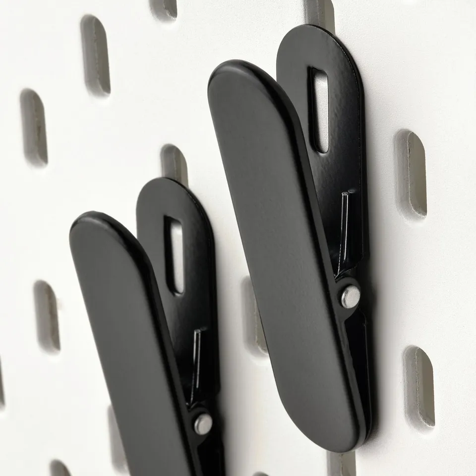 Black metal hooks on a white pegboard. Sturdy clips, arranged neatly, for organising items.