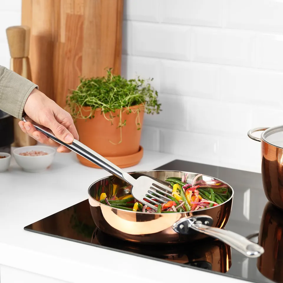 Hand holds FINMAT fork stirring mixed salad in shiny copper bowl on induction stove, near herbs.