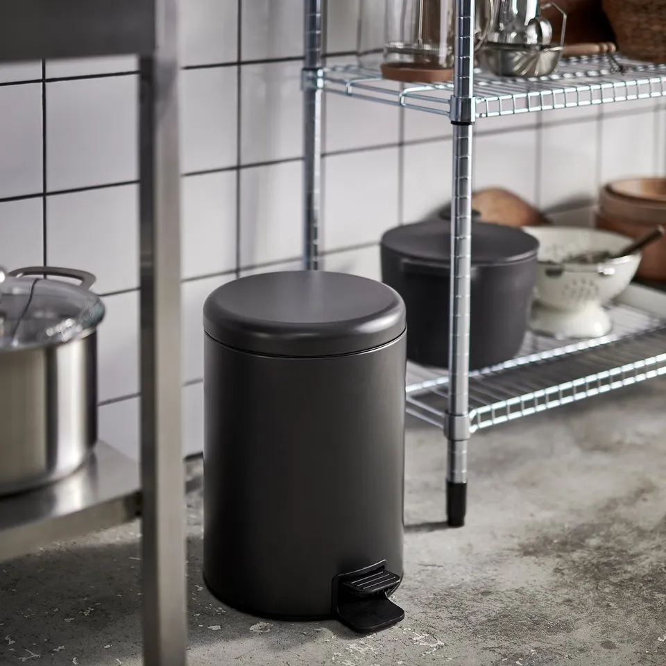 Small black pedestal bin in kitchen with wire shelves, pots, and bowls.