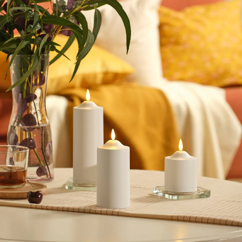 Three white SMÅLOM led candles on a table, surrounded by a vase with flowers and a glass with cherries. Candles are smaller than the vase.