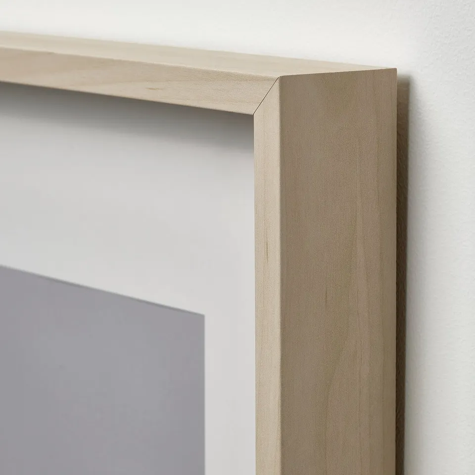 Wooden picture frame with light finish and plastic front protection, leaning against a white wall with a grey picture partially visible inside.