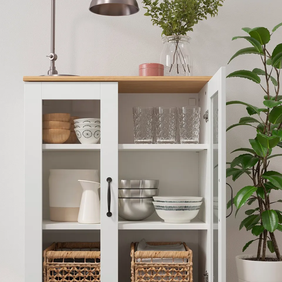 Kitchen cabinet with glass doors showing bowls, glasses, baskets, and a plant on the counter.