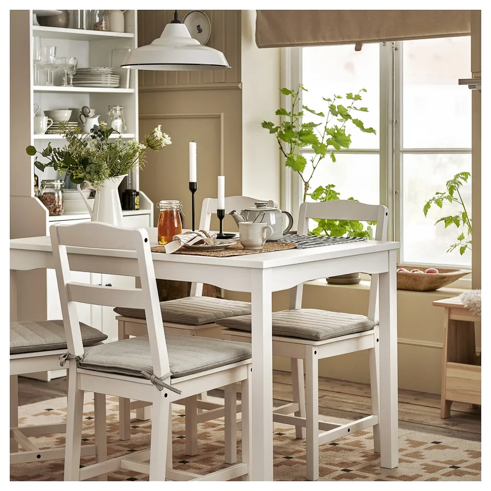 Cosy dining area with flowers, candlesticks, and teapot. Sunlight streams in from a window with dishes on shelves.