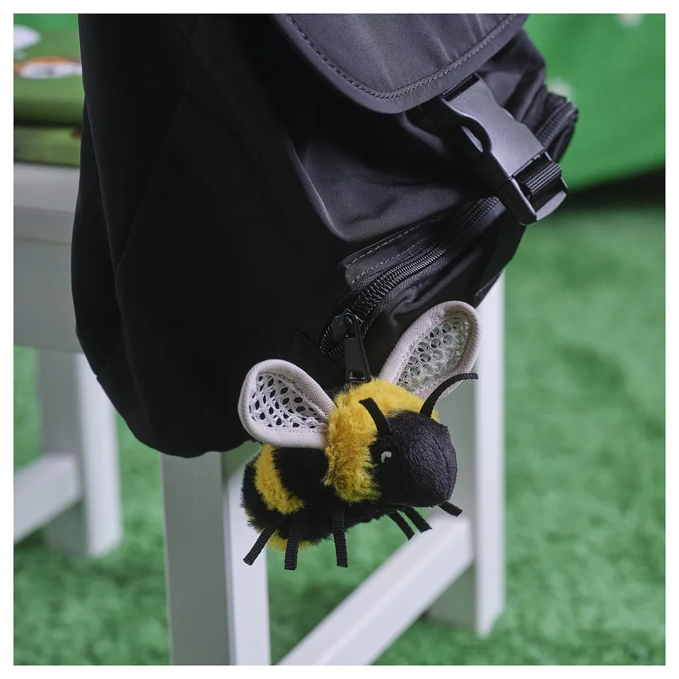 A black backpack with a plush bee toy attached. The bee is yellow and black, with white mesh wings and it's looped through a zip pull.