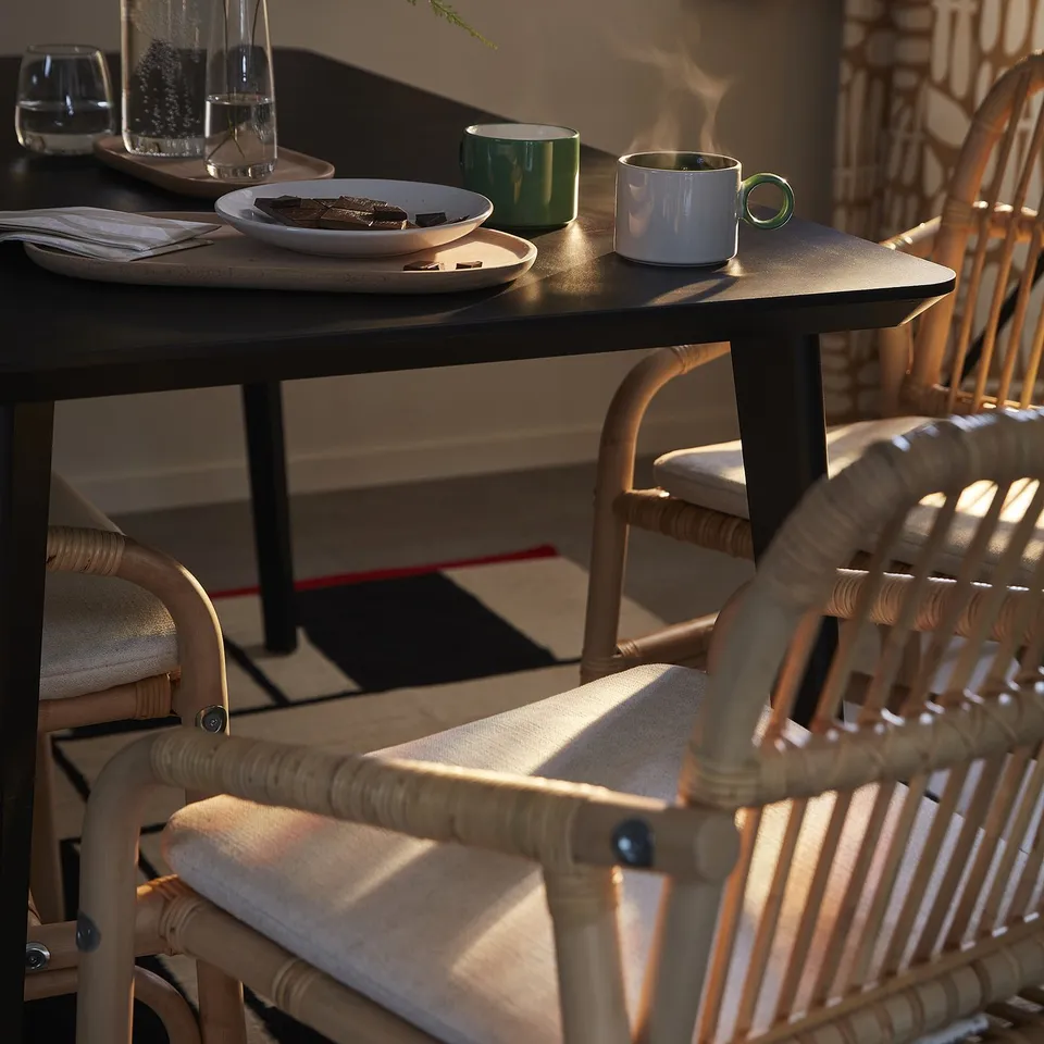 Rattan chair with cushion, modern traditional design, beside table with food and mugs, near window with sunlight.