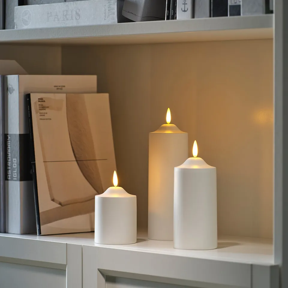 Three glowing SMÅLOM led candles in sizes on shelf, flickering, surrounded by books.