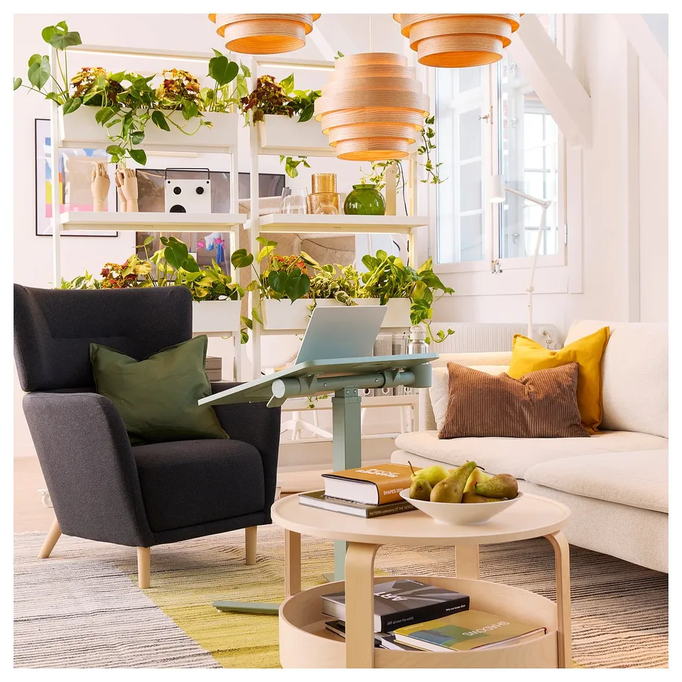 Modern living space with grey armchair, white laptop table, and vibrant decor. Shelves filled with plants, books, and decorative items.