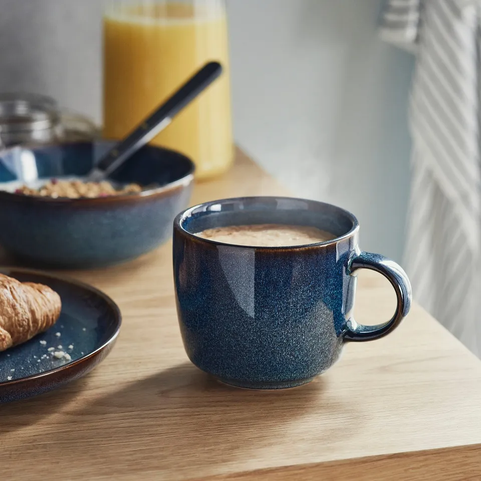 Blue mug filled with coffee on a wooden table, next to a bowl of cereal and pastry.