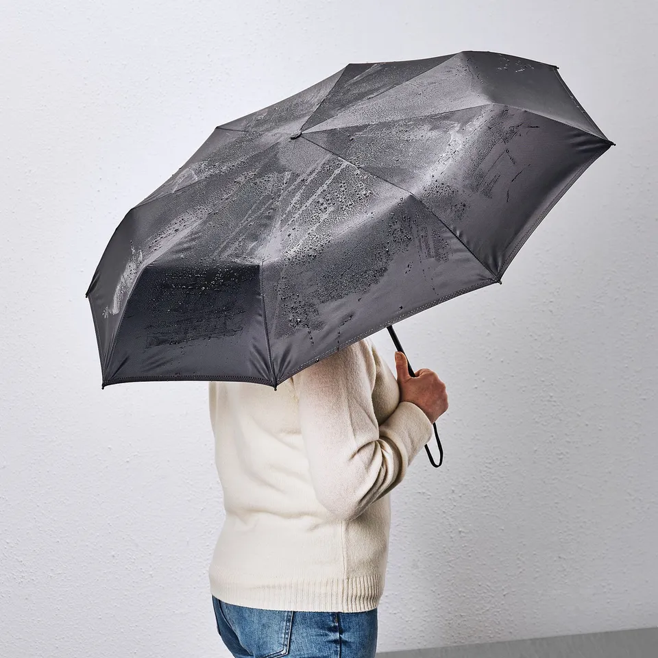 A person holding a black KNALLA umbrella, which is foldable and provides protection from heavy rain. The handle has a strap for easy carrying.