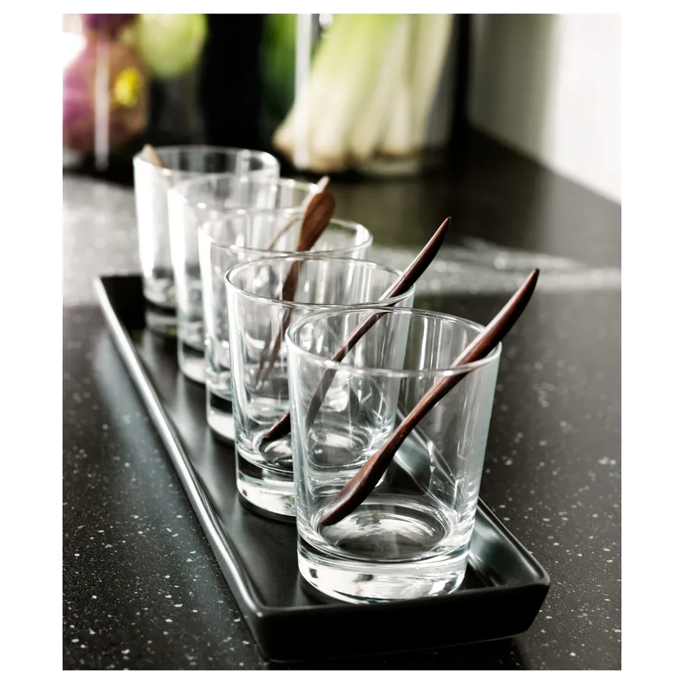Six clear glasses on black tray, each with a wooden stirrer, set on dark speckled countertop.