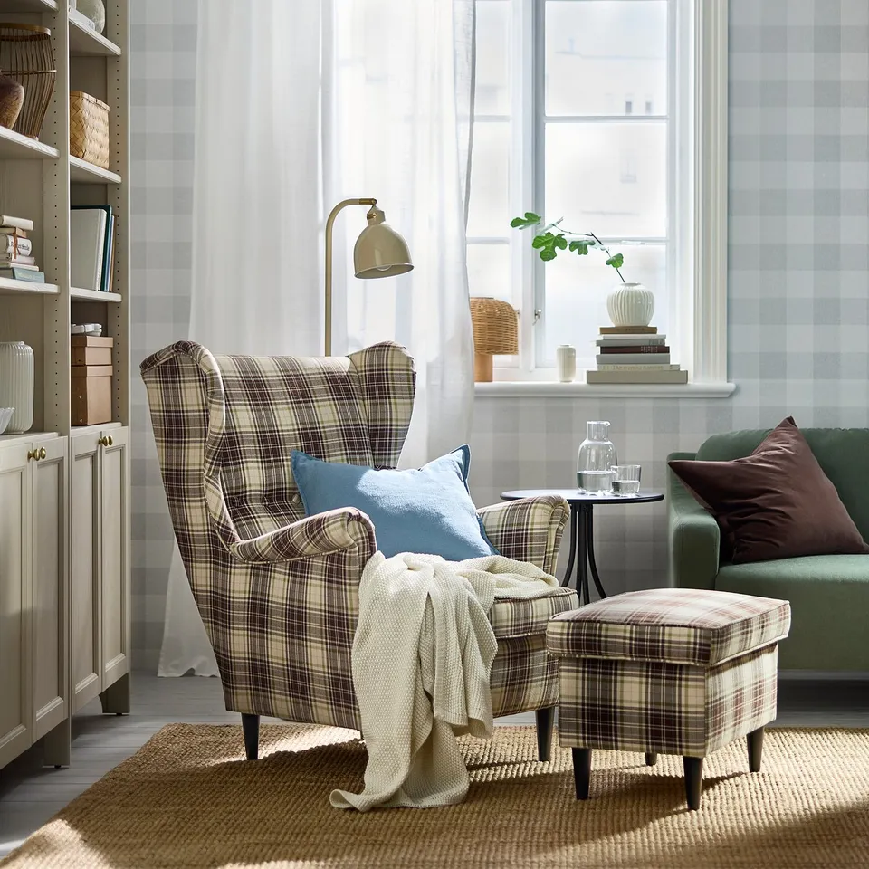 Cosy living room with STRANDMON wingback chair, matching footstool, green sofa, large window, and wooden shelves. Warm, inviting decor.