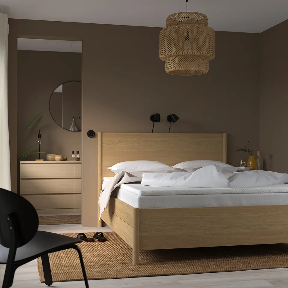Cosy bedroom with wooden bed, white linens, and pendant light. NORDBERGET pad, black chair, beige walls, lamp, minimal decor.