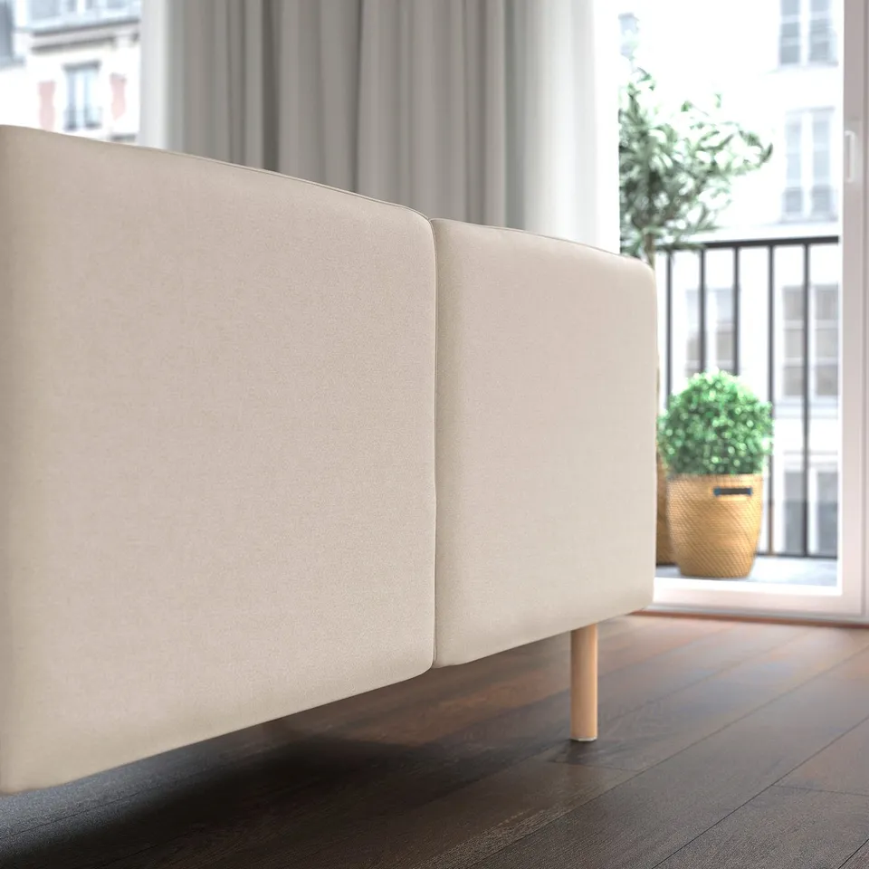 A modern beige sofa facing a window with a plant. The sofa shows clean lines and wooden legs.
