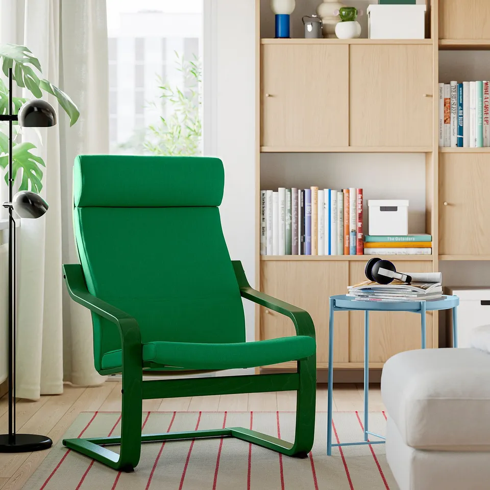 Modern living room with green nasa POÄNG armchair, padded with green fabric, next to blue side table and wooden bookshelf, near window with plant.