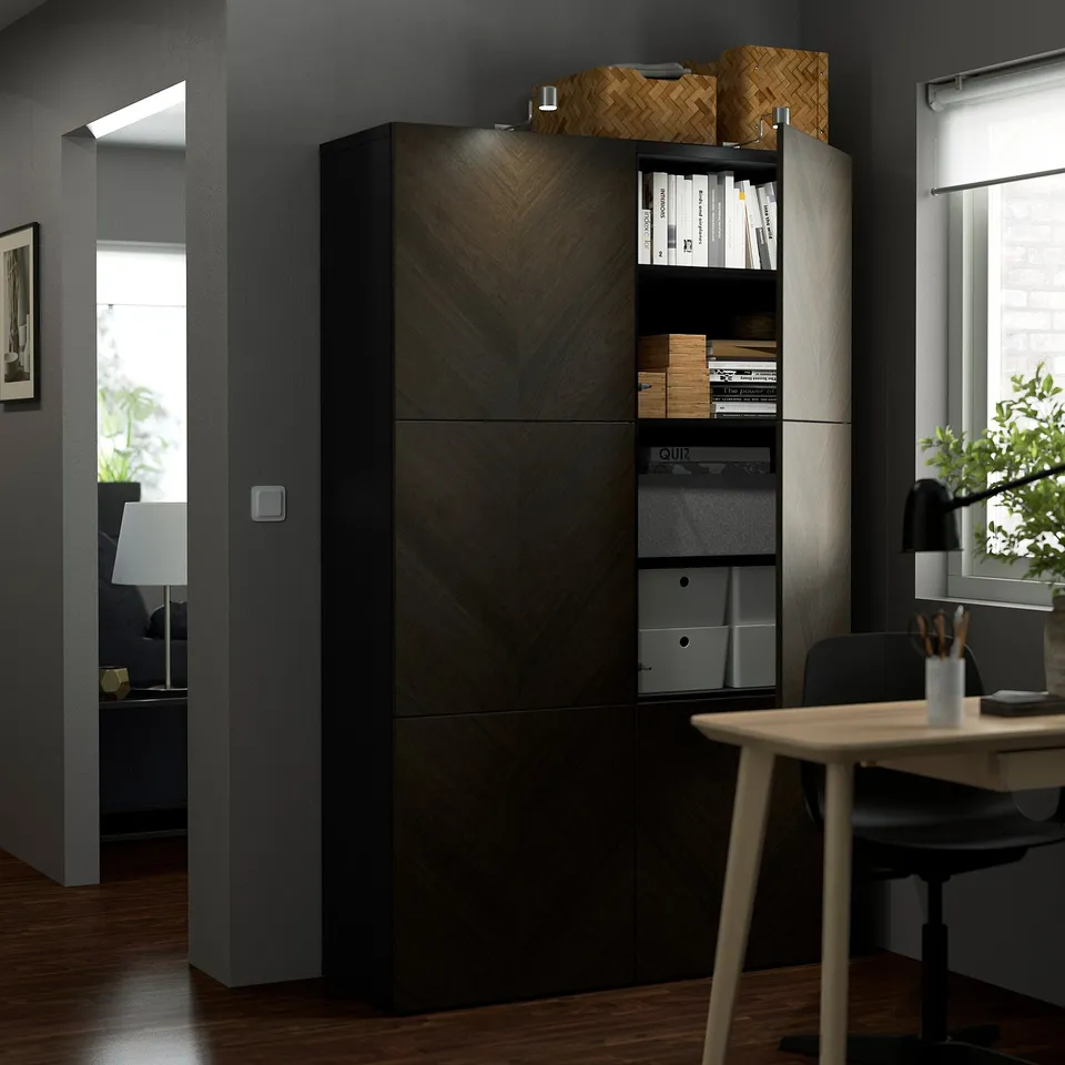 Modern home office with wooden BESTÅ shelves, holding files and books. A desk with a chair and lamp is beside it.
