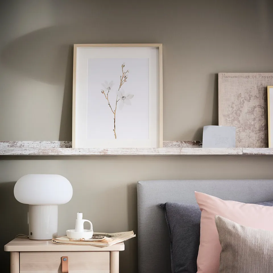 Modern bedroom with light wood bedside table, white DEJSA lamp, grey and pink pillows, framed floral art, and a floating shelf.