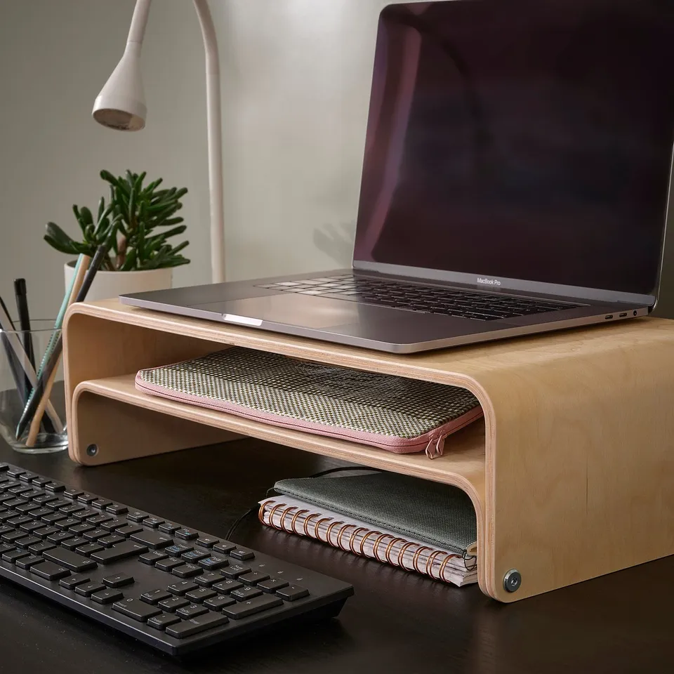 A modern workspace with a ROMELSÖN laptop stand in natural wood, holding a purple-screen laptop. A black keyboard, plant, and desk lamp add ambiance.