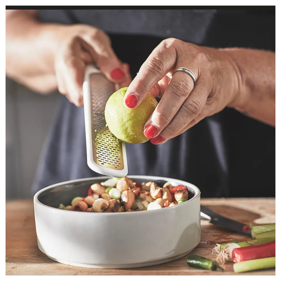Person grates lime zest into bowl of mixed nuts; white grater, red nails, and knife nearby.