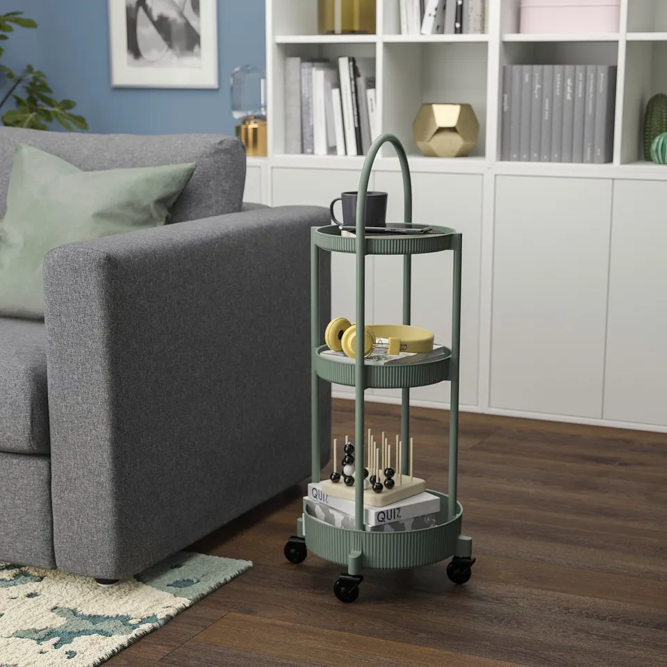 Small green cart next to grey couch, holding cups, books, and decor, positioned between floor rug and wooden shelf.