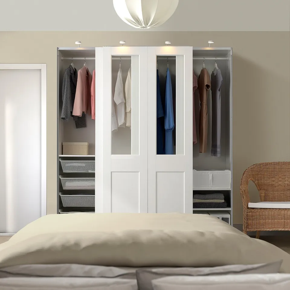 White PAX wardrobe with glass doors, clothes, and baskets. Beige bed and wicker chair nearby. Dominates room.