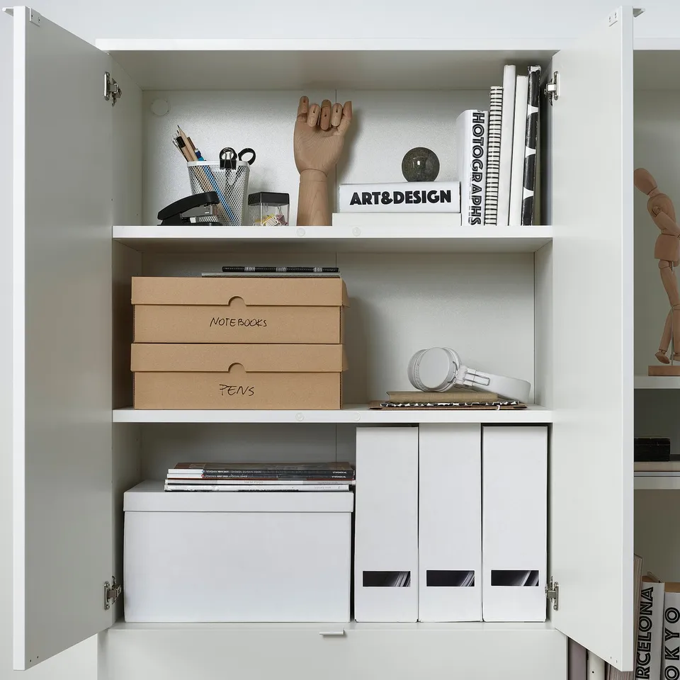 White cabinet with shelves storing stationery, art books, and cardboard boxes labelled notebooks and pens for organised storage.