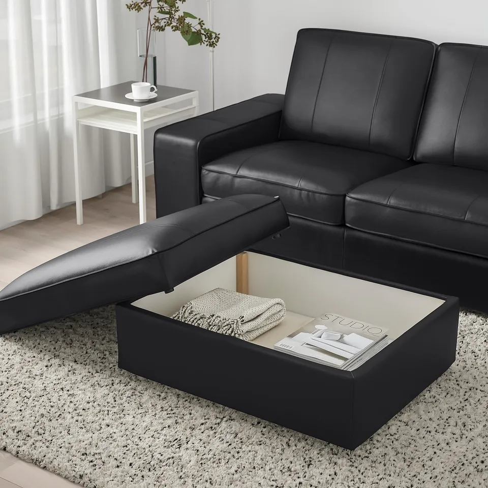 Modern black leather sofa with footstool featuring hidden storage, open showing blanket and magazine. White rug and side table with cup.
