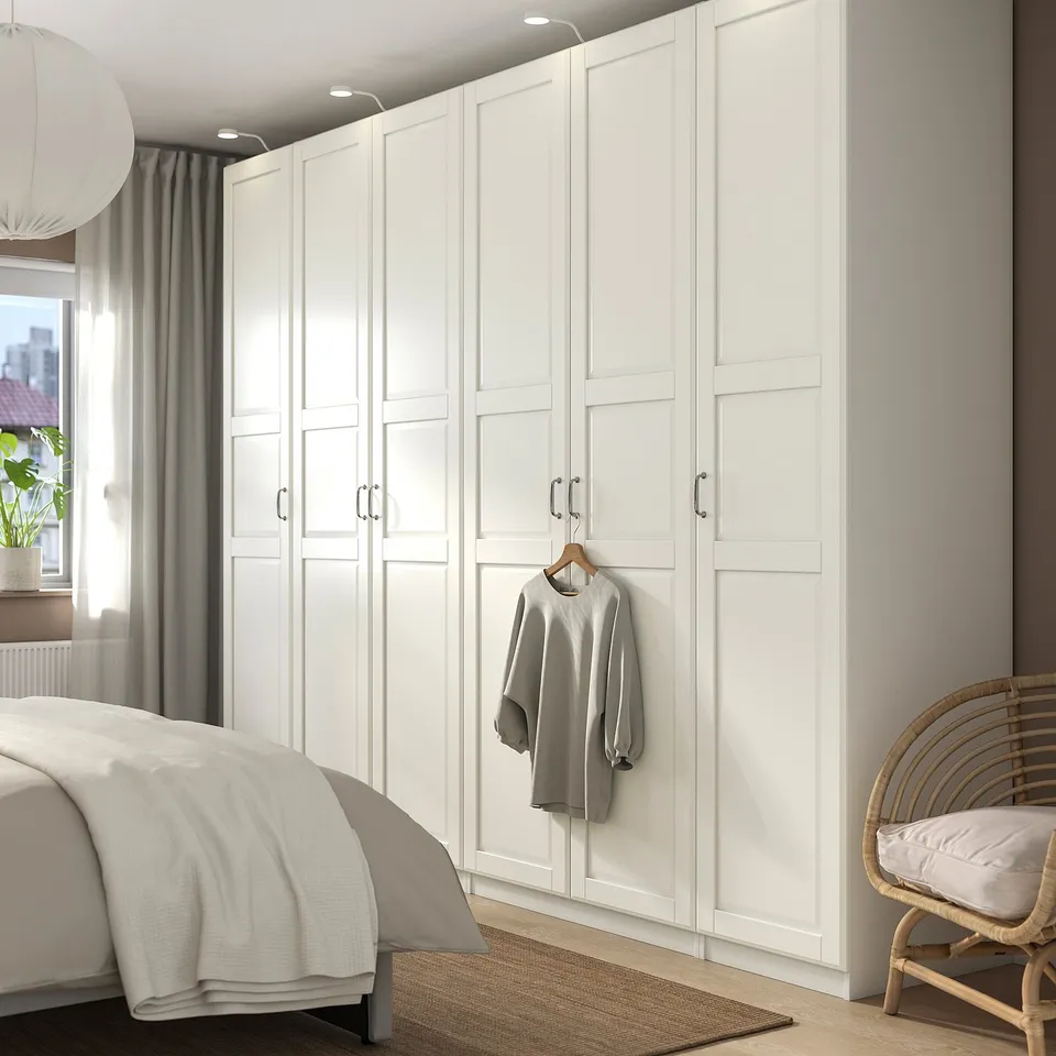 Modern white floor-to-ceiling PAX wardrobe. Tall, sleek design dominates room, housing grey sweater. Nearby, white bed, woven chair.