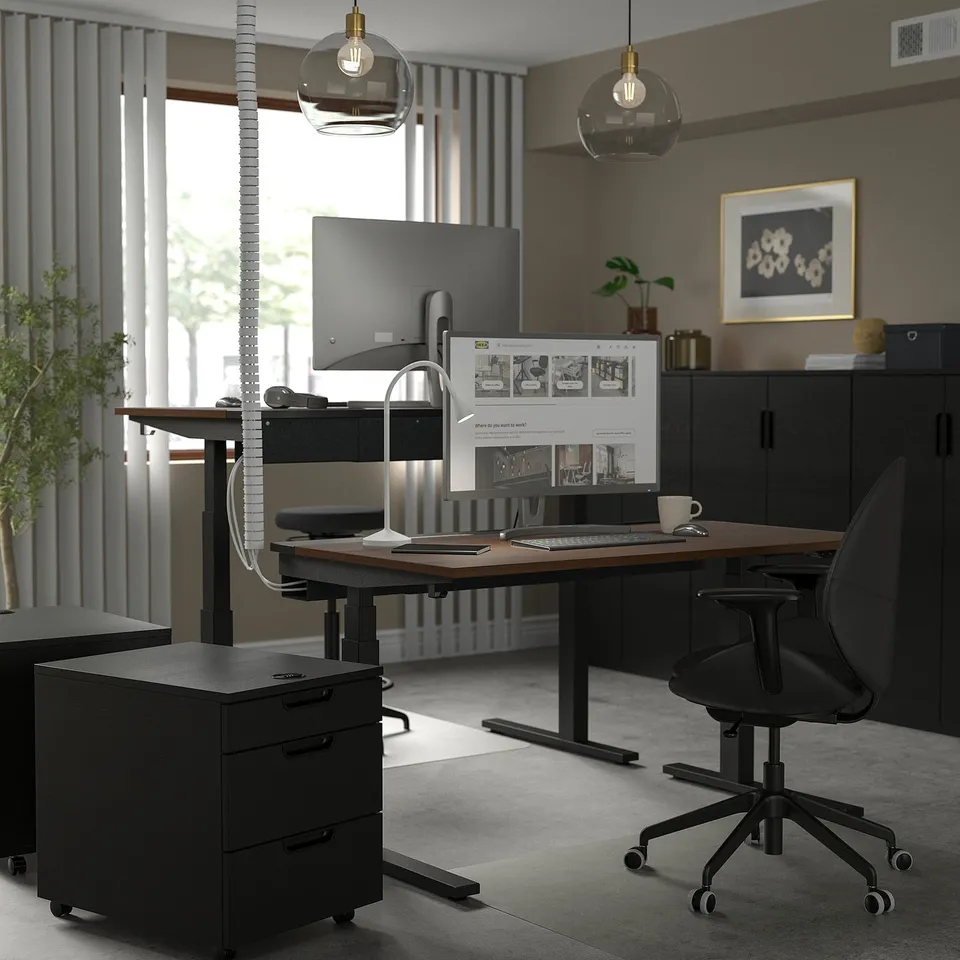 Modern home office with MITTZON black sit-stand desk, ergonomic chair, dual monitors, laptop, and pendant lights. Open floor plan, light-filled space.