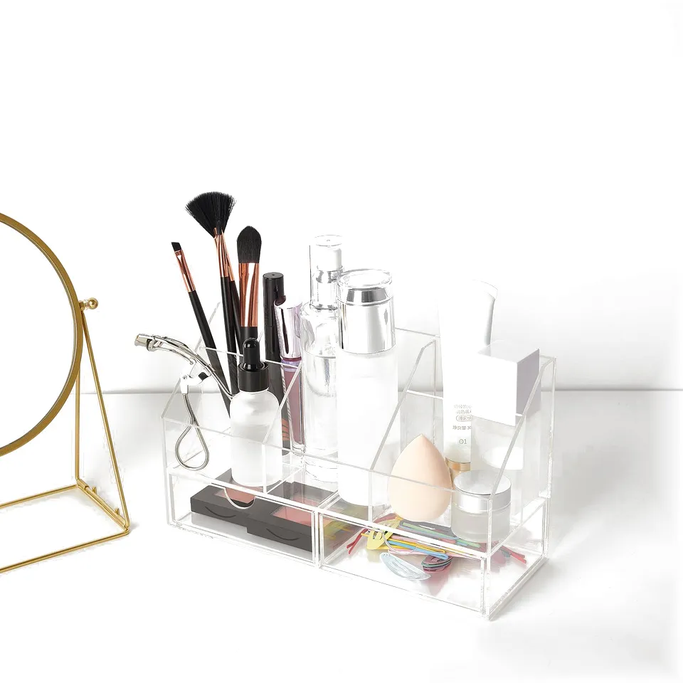 A clear acrylic organiser holds makeup brushes, bottles, and accessories neatly on a white surface next to a gold mirror.