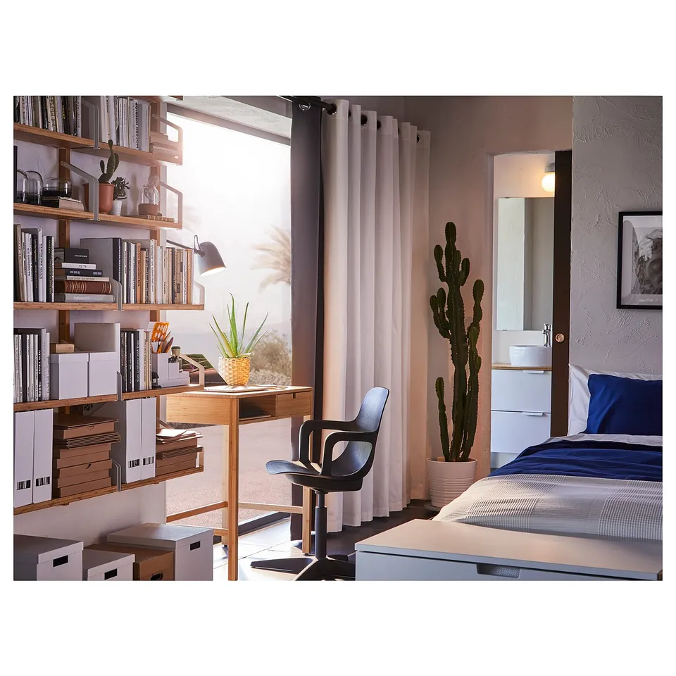 Bedroom with wooden desk, chair, shelves, and bed. Blue bed linens and large cactus plant. Curtains and natural light from window.