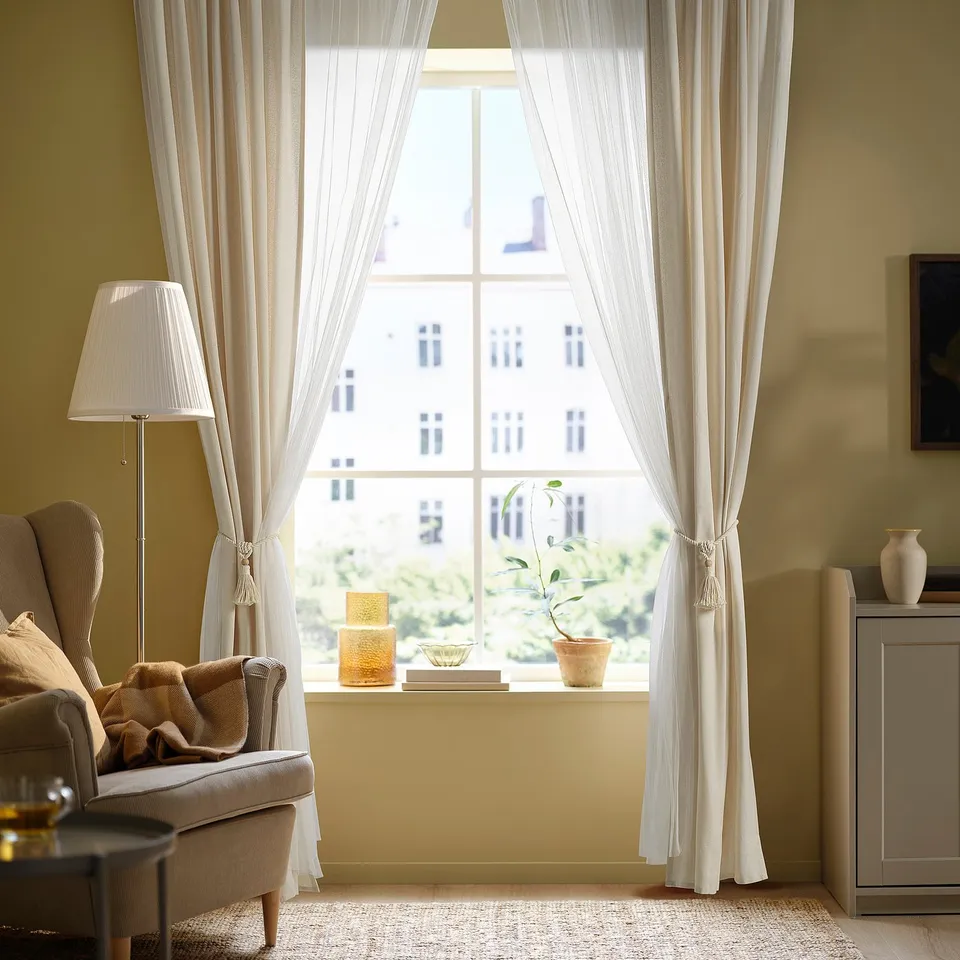 Cosy living room with DUNFLÄTA tie-backs, sheer curtains, and yellow building view. Beige armchair and lamp.