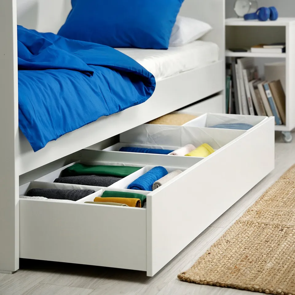 Open under-bed storage with color-coded folded clothes in white dividers.
