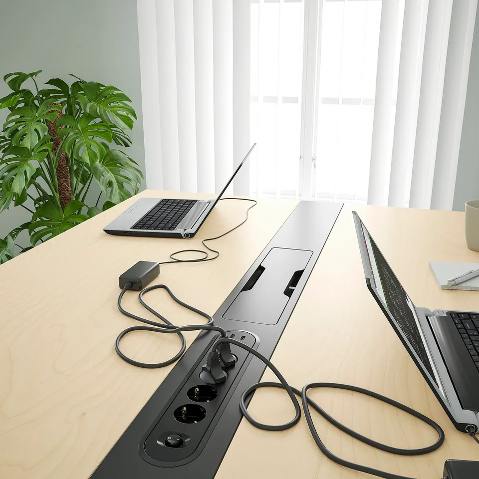 Modern conference table with power strips, laptops, and plants. Cable management module included for charging devices.