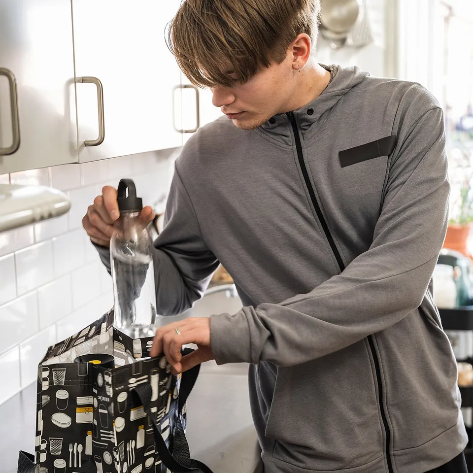 Person packs plastic bottle in grey FLADDRIG lunch bag with kitchen utensil design.