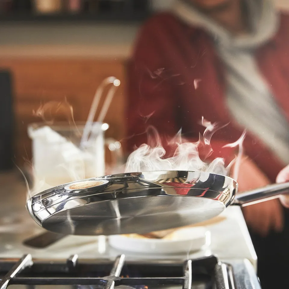 A person cooks with a stainless steel frying pan on a stove, steam rising, showcasing its durability and even heat distribution.