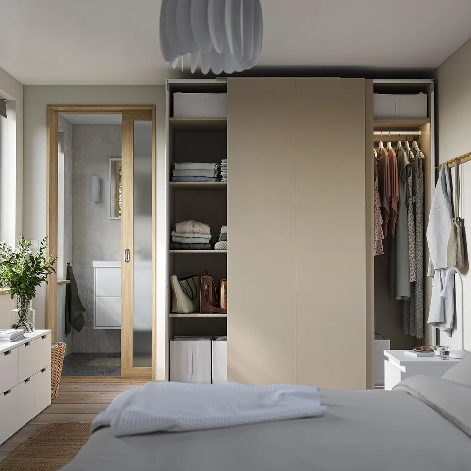 Modern bedroom with beige PAX wardrobe, open shelves, and neat clothes. White bed, pendant lamp, and bathroom entrance visible.